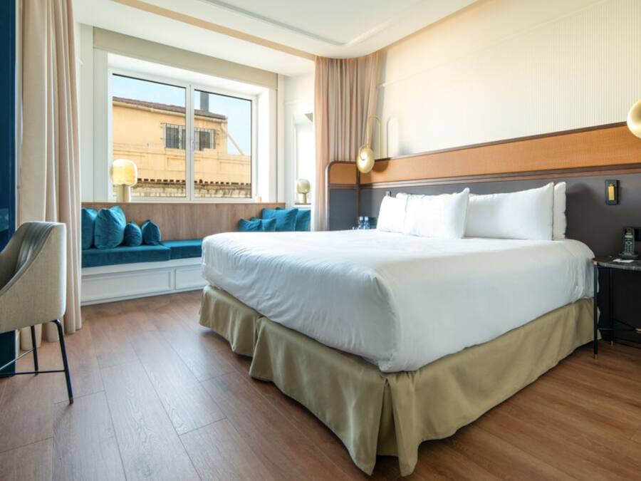 Habitación de hotel con una cama grande, un escritorio con silla, una ventana con vistas a edificios y una mesita de noche con lámpara.