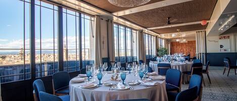 A restaurant with round tables set for dining, large windows offering a view, and a modern chandelier.