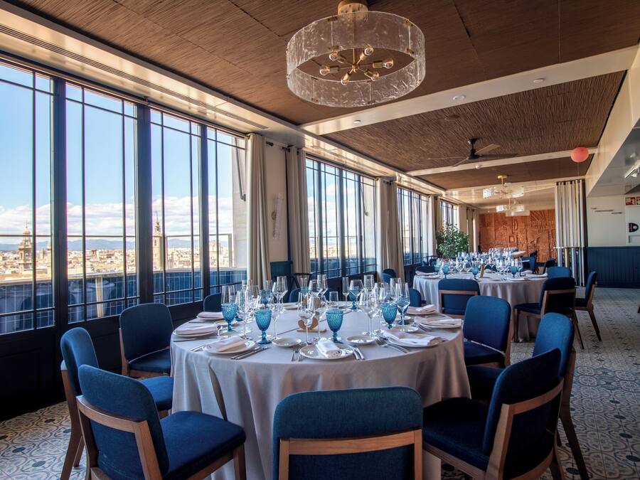 Un restaurante con mesas redondas dispuestas para comer, ventanas amplias que ofrecen vistas y una moderna lámpara de araña.