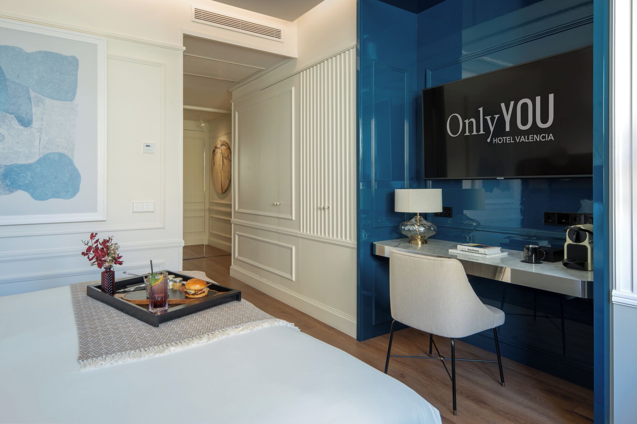 A hotel room with a bed, a desk, a chair, a TV, and a tray with food and drink.