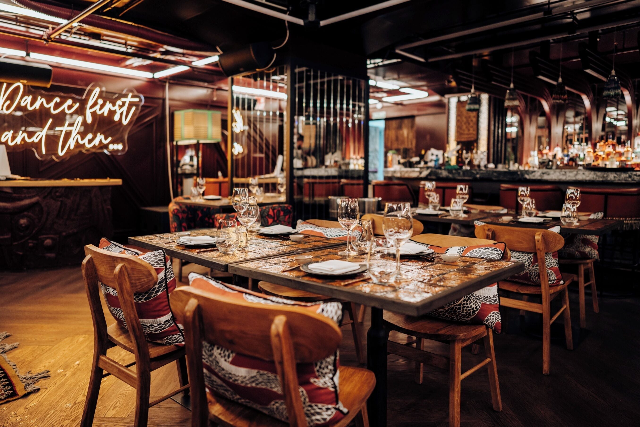 A restaurant interior with multiple tables set for dining, chairs with patterned cushions, and a bar area in the background.