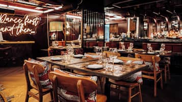 A restaurant interior with multiple tables set for dining, chairs with patterned cushions, and a bar area in the background.