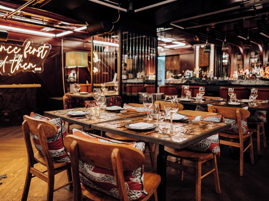 Interior de restaurante con varias mesas dispuestas para comer, sillas con cojines estampados y una zona de bar al fondo.