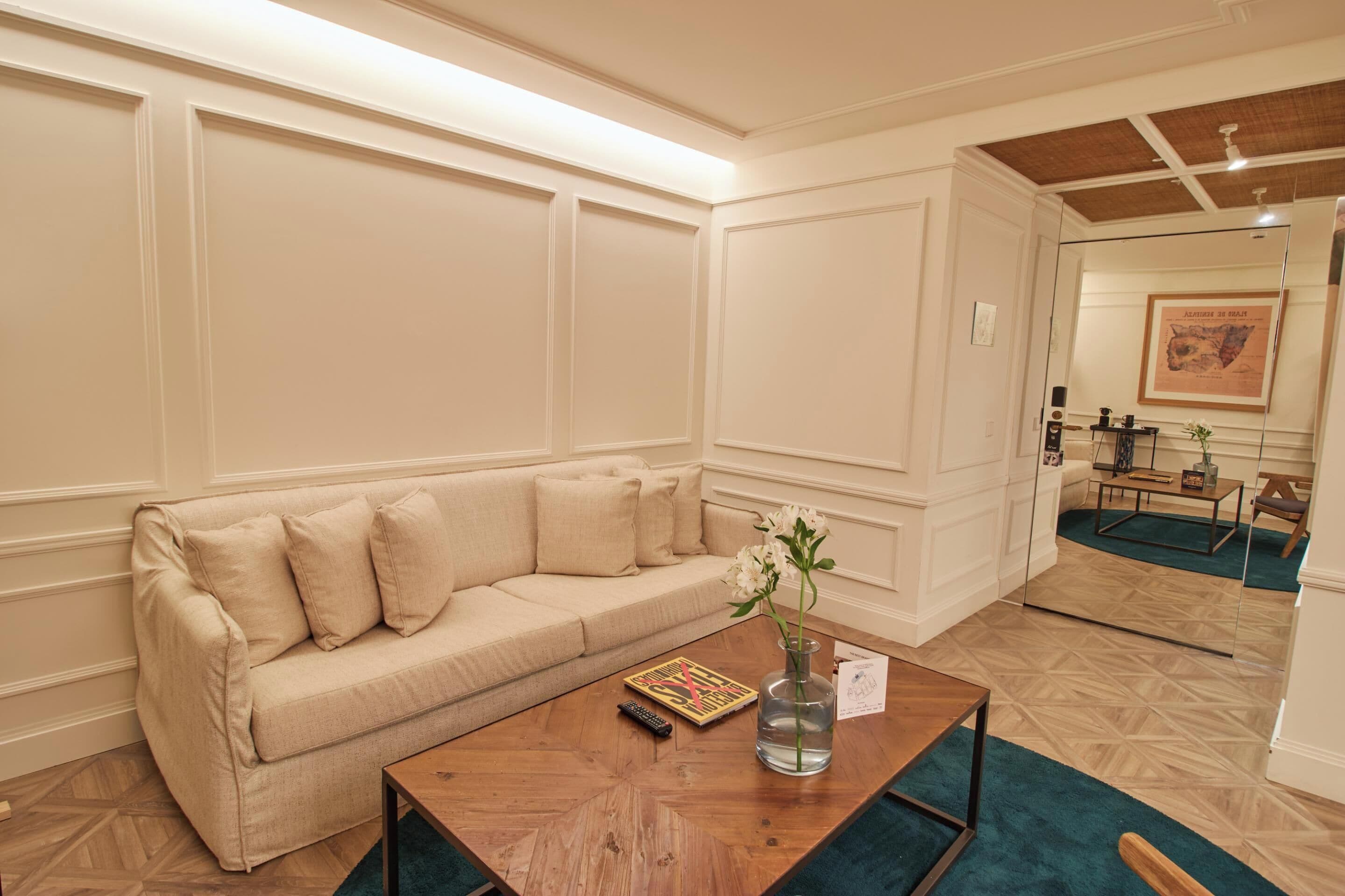 Una habitación con un sofá color beige, una mesa de centro de madera con un jarrón de flores y un cuadro en la pared.