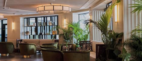 A lobby with a modern design, featuring a seating area with green upholstered chairs, a large potted plant, and a decorative wall with shelves.