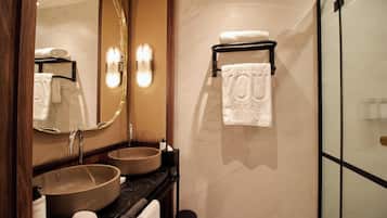 Un baño con un espejo grande, dos lavamanos, un perchero con toallas y una cabina de ducha con mampara de vidrio.