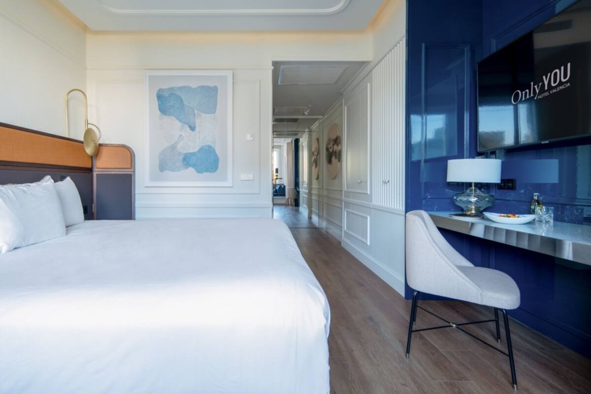 A hotel room with a large bed, a desk with a chair, a painting on the wall, and a view of the hallway.