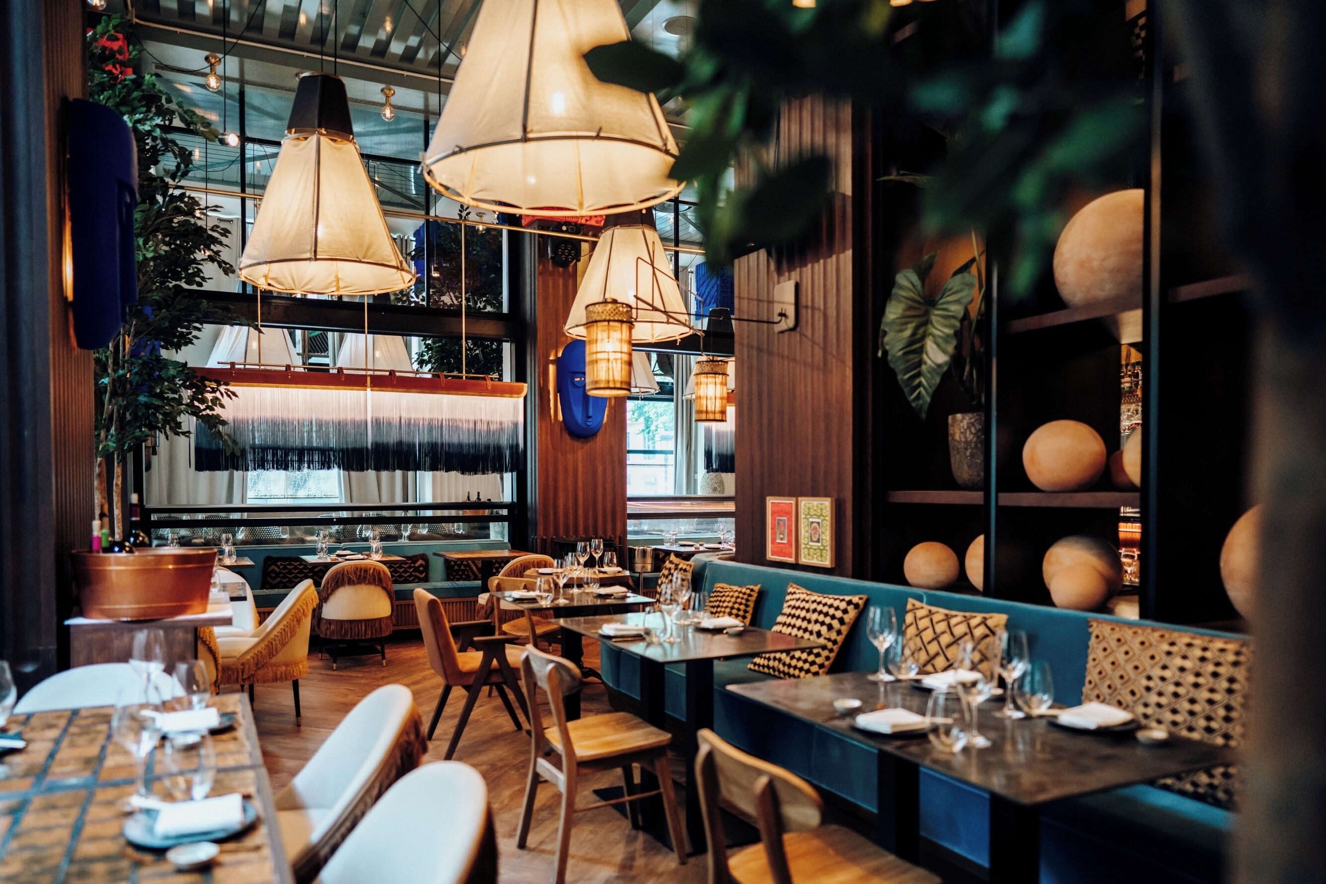 A restaurant interior with wooden tables, cushioned seating, hanging pendant lights, and a view through large windows.