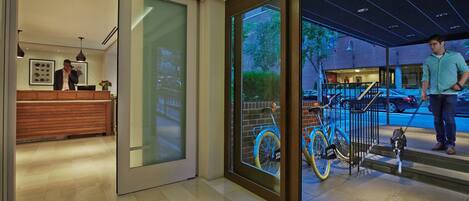 A hotel reception area with a wooden counter, a receptionist, and a glass door leading outside.