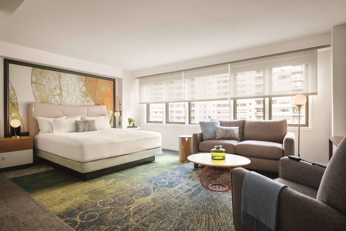 Una habitación de hotel moderna con una cama grande, un sofá, una mesita de café y vistas a la ciudad a través de amplios ventanales.
