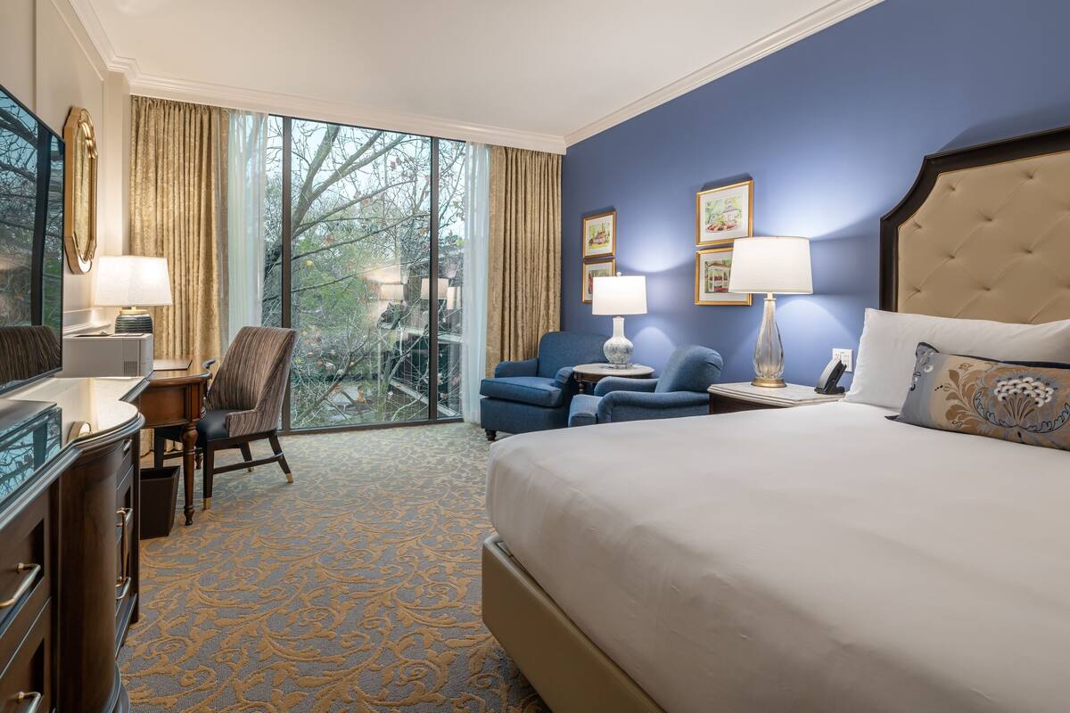 Une chambre d’hôtel avec un grand lit, un bureau avec une lampe, deux fauteuils, un canapé et une vue sur les arbres à travers de grandes fenêtres.