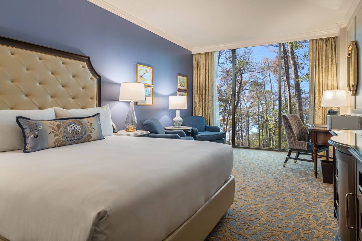 Une chambre d’hôtel avec un grand lit, deux fauteuils, un bureau et une vue sur les arbres par la fenêtre.