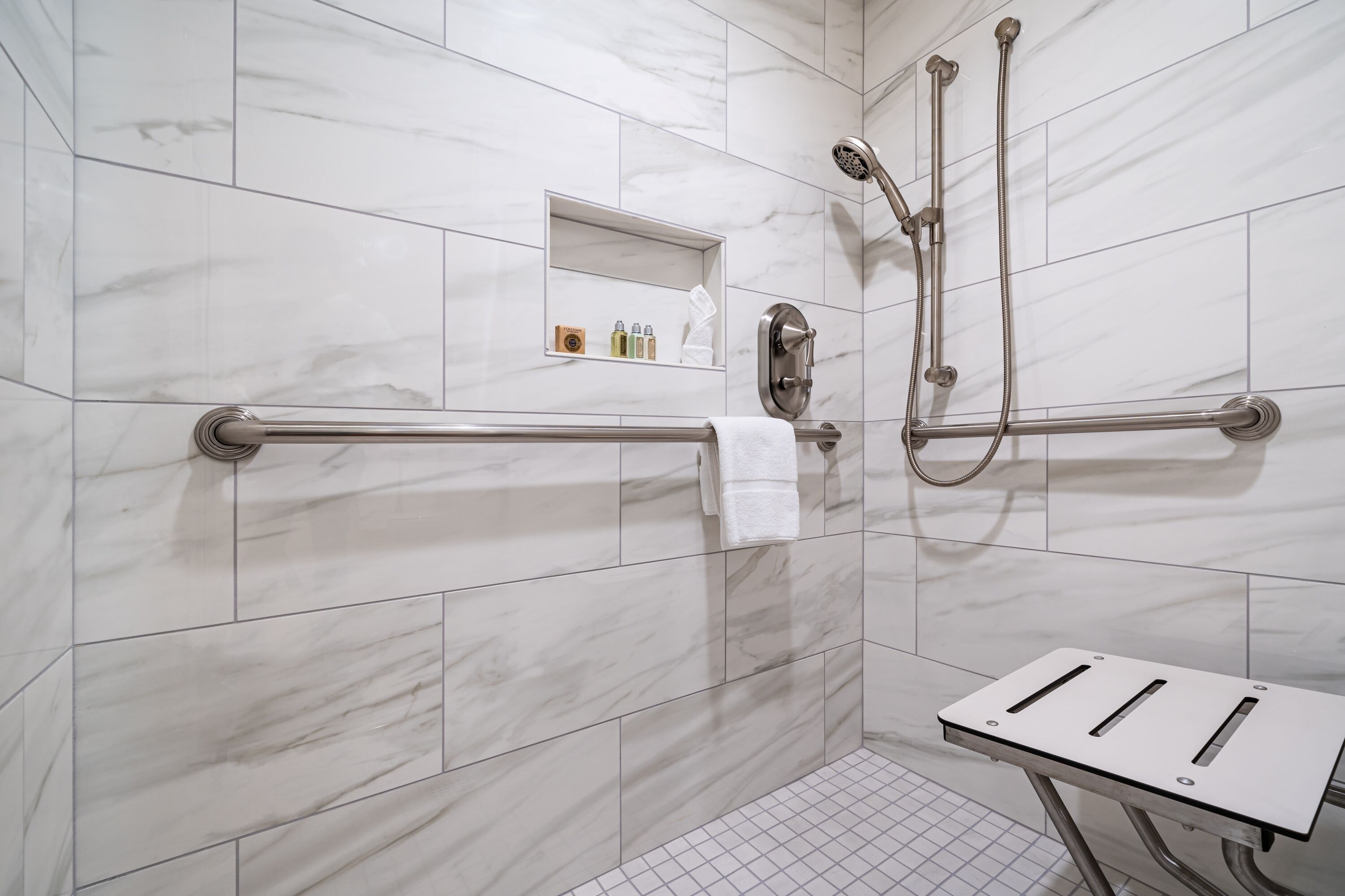 Une salle de bain carrelée de marbre blanc, une douche avec un pommeau de douche amovible, un porte-serviettes et une petite étagère contenant des articles de toilette.