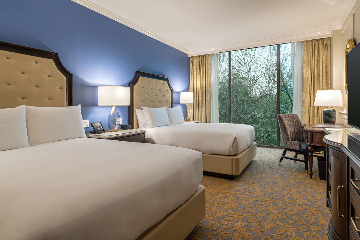 Une chambre d’hôtel avec deux lits, un bureau, une chaise et une grande fenêtre donnant sur les arbres.
