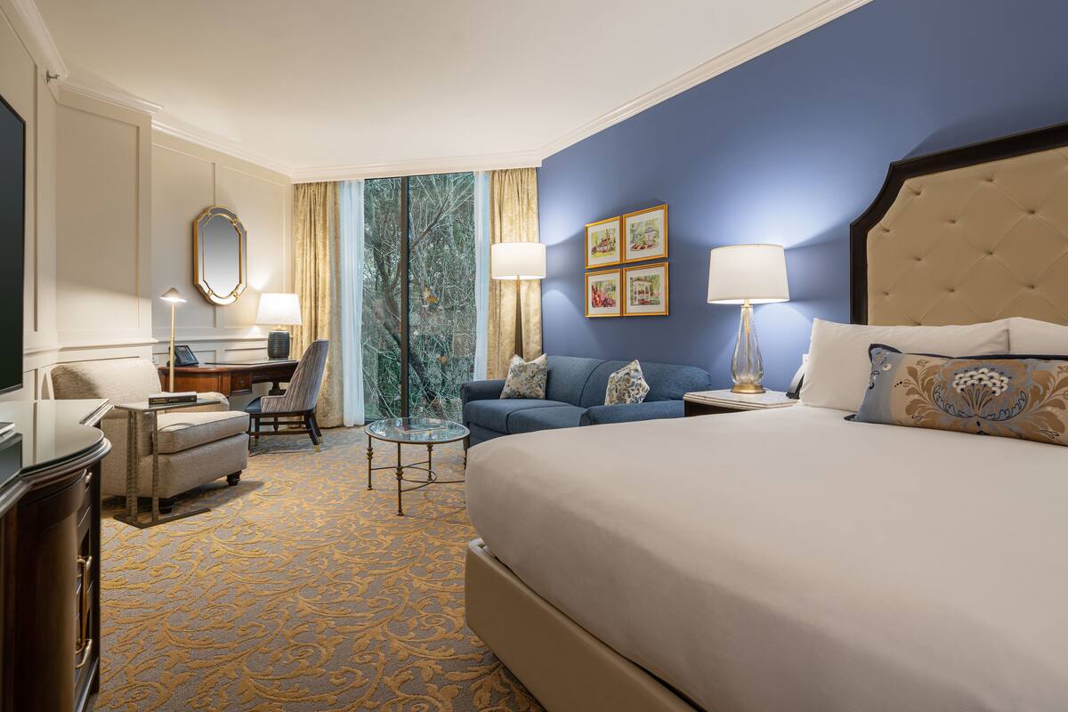 Une chambre d’hôtel avec un grand lit, un canapé, un bureau et une chaise. Il y a une fenêtre avec des rideaux, une lampe et des tableaux encadrés au mur.