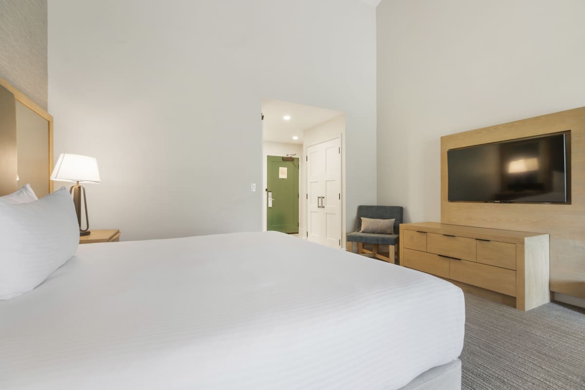 Una habitación de hotel con una cama grande, un televisor montado en la pared, un escritorio con cajones, una silla y una lámpara en una mesita de noche.