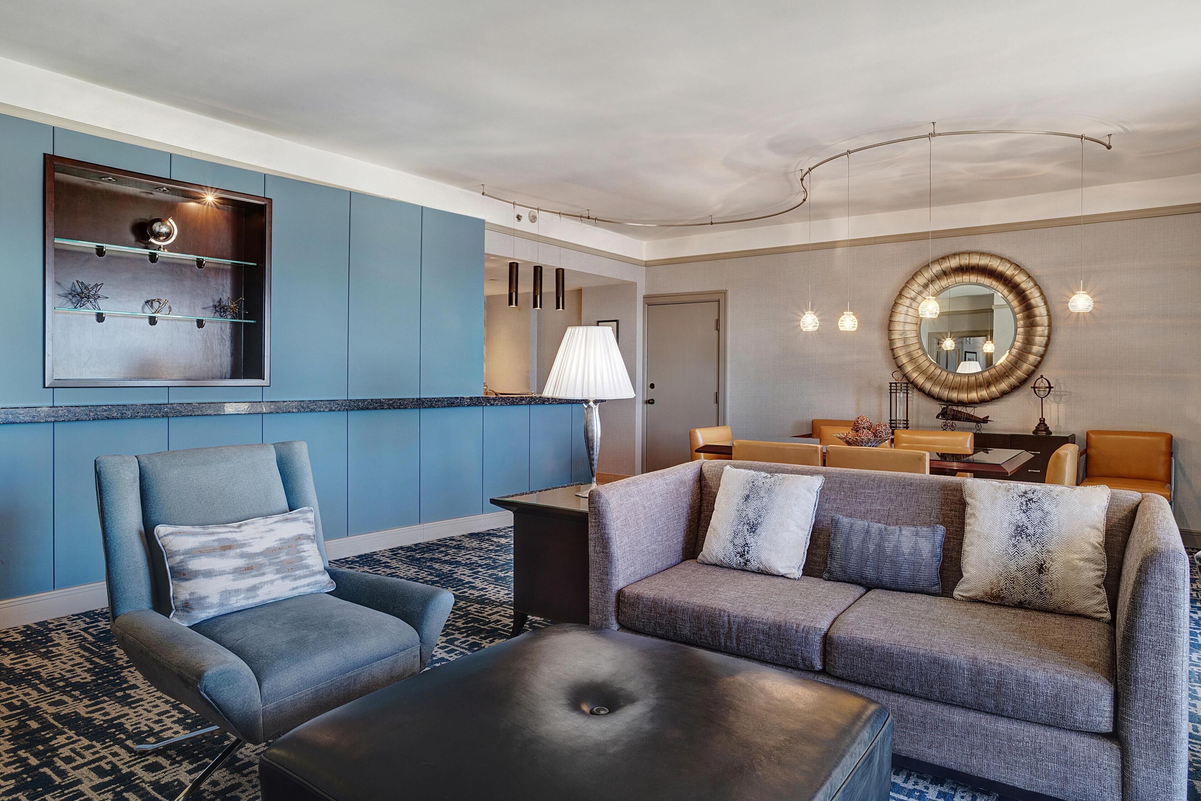 En moderne hotellobby med en blå accentvæg, et siddeområde med sofa og lænestole, et rundt spejl og et lille bord med en lampe.