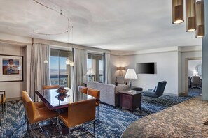 A hotel room with a dining table, chairs, a sofa, a TV, and a large window with curtains.
