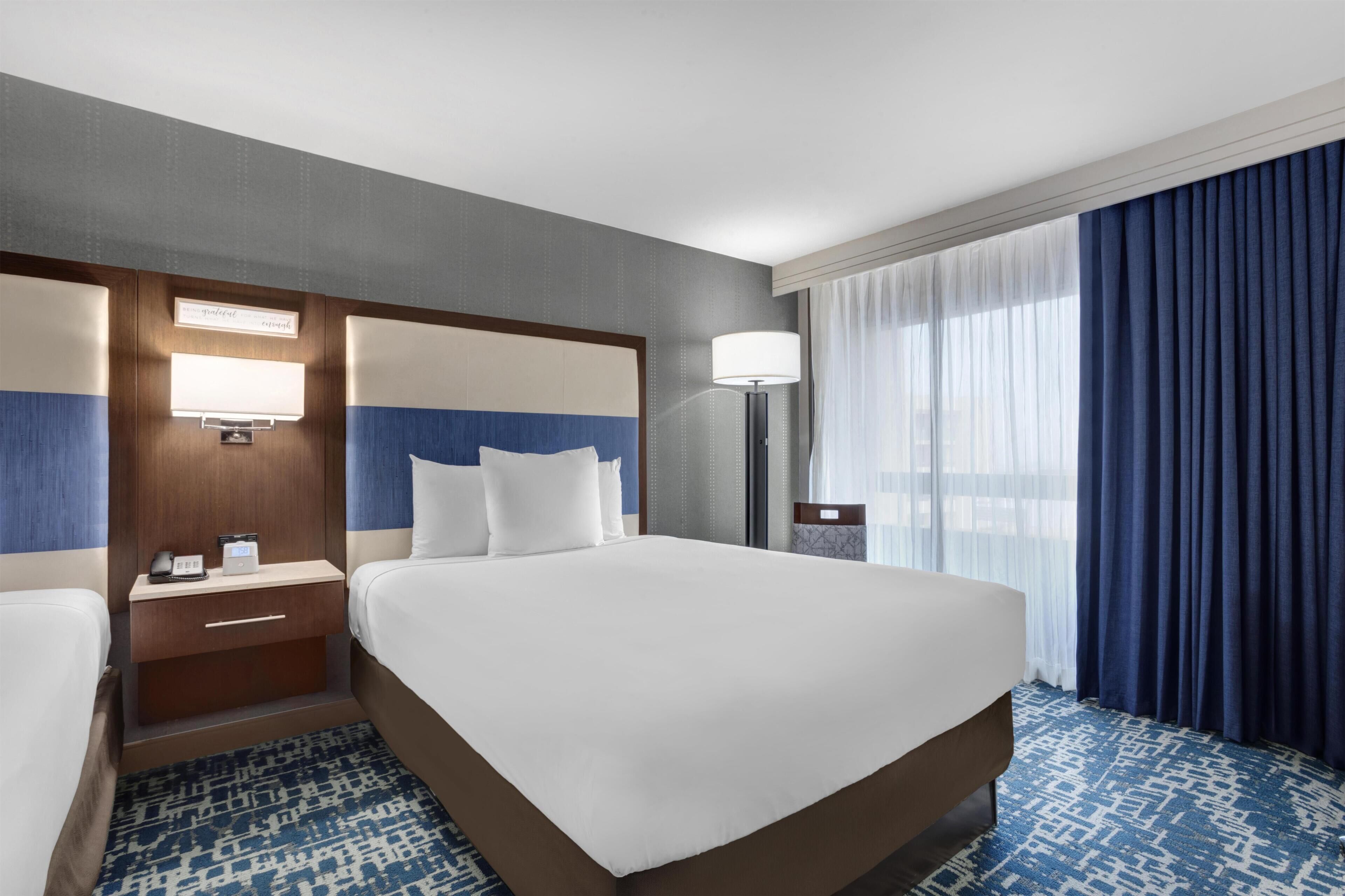 Habitación de hotel con una cama grande, una mesita de noche con lámpara, un escritorio con teléfono y una ventana con cortinas.