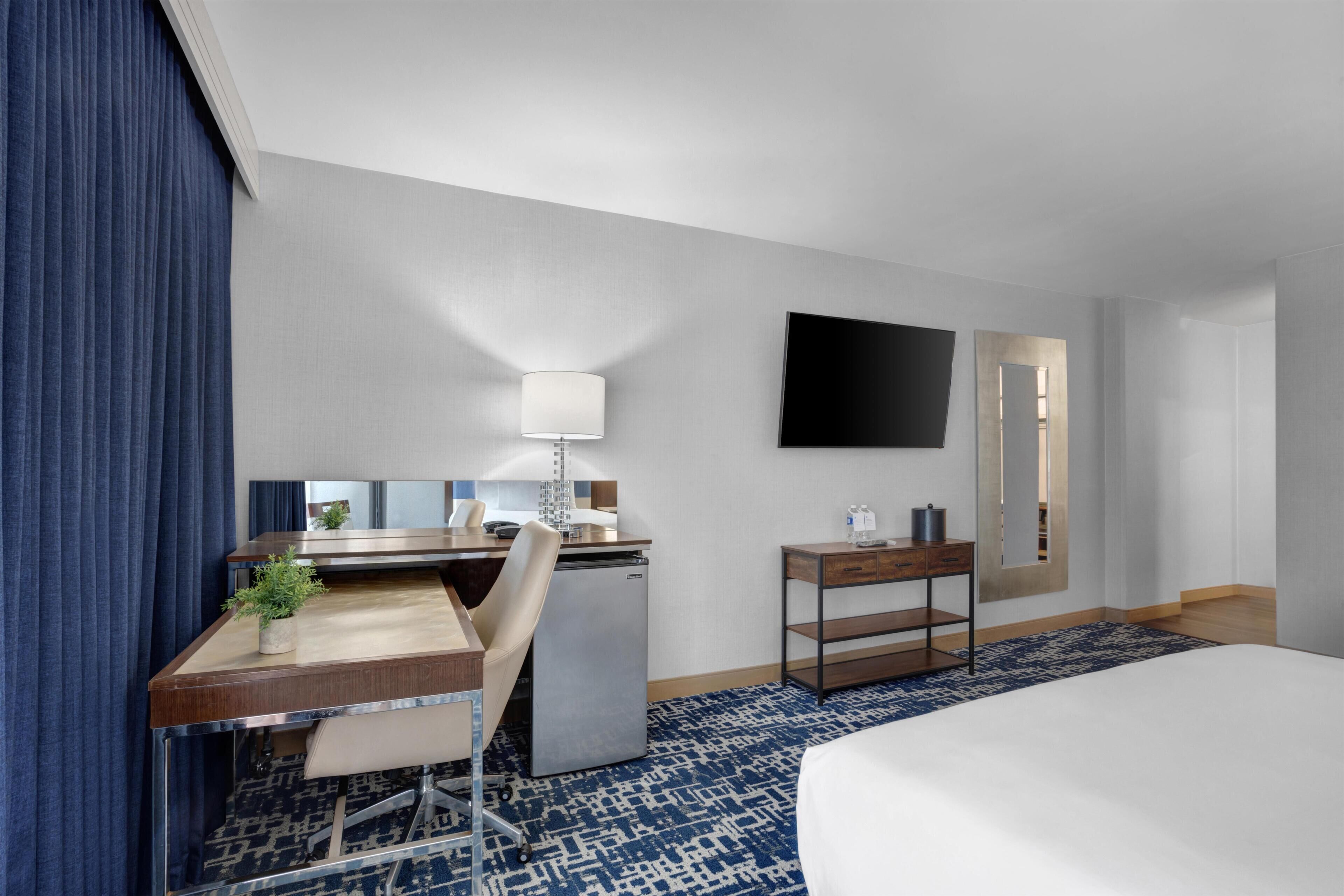 A hotel room with a bed, desk, chair, lamp, TV, and a small cabinet.