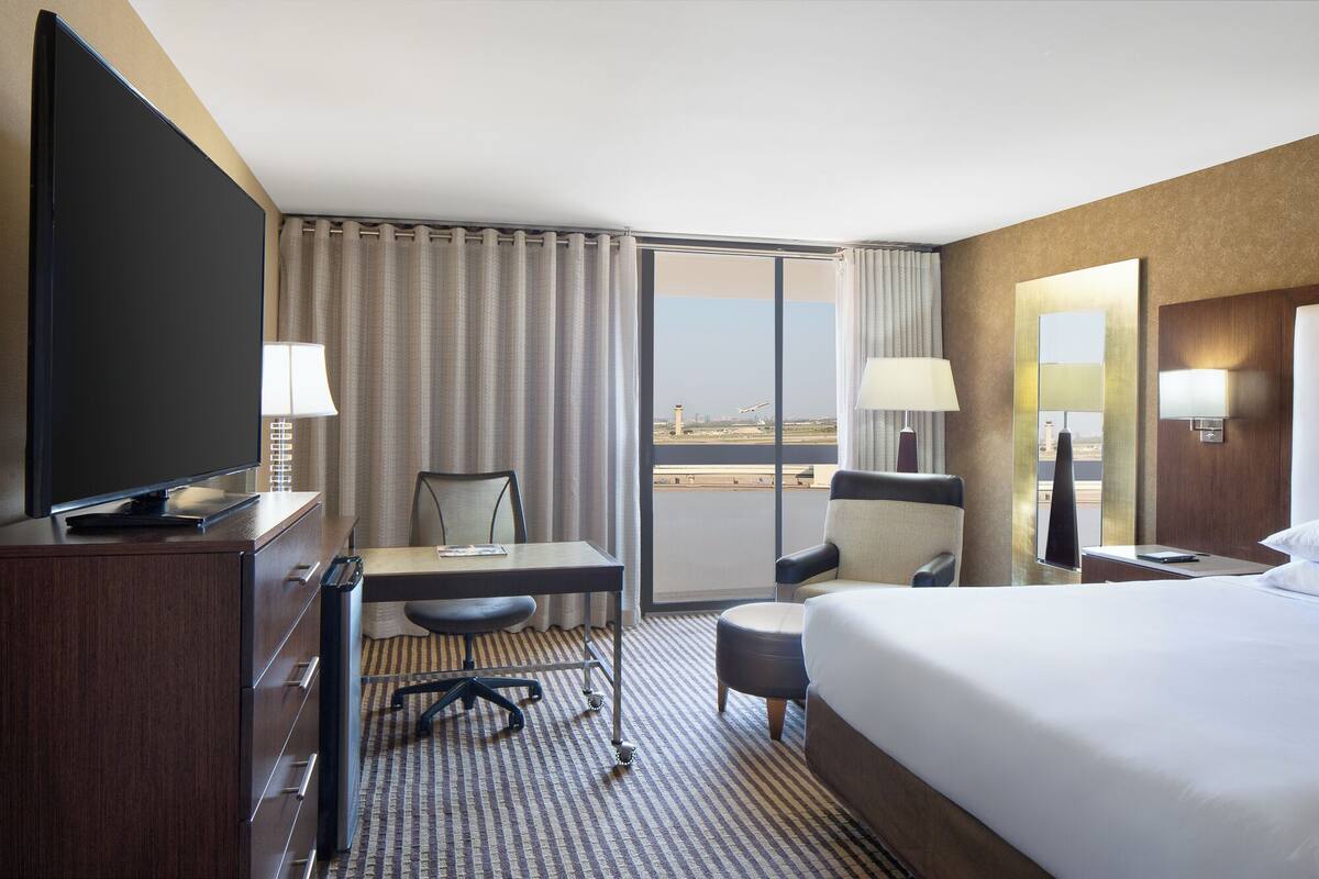 A hotel room with a bed, desk, chair, TV, and a view of the airport.