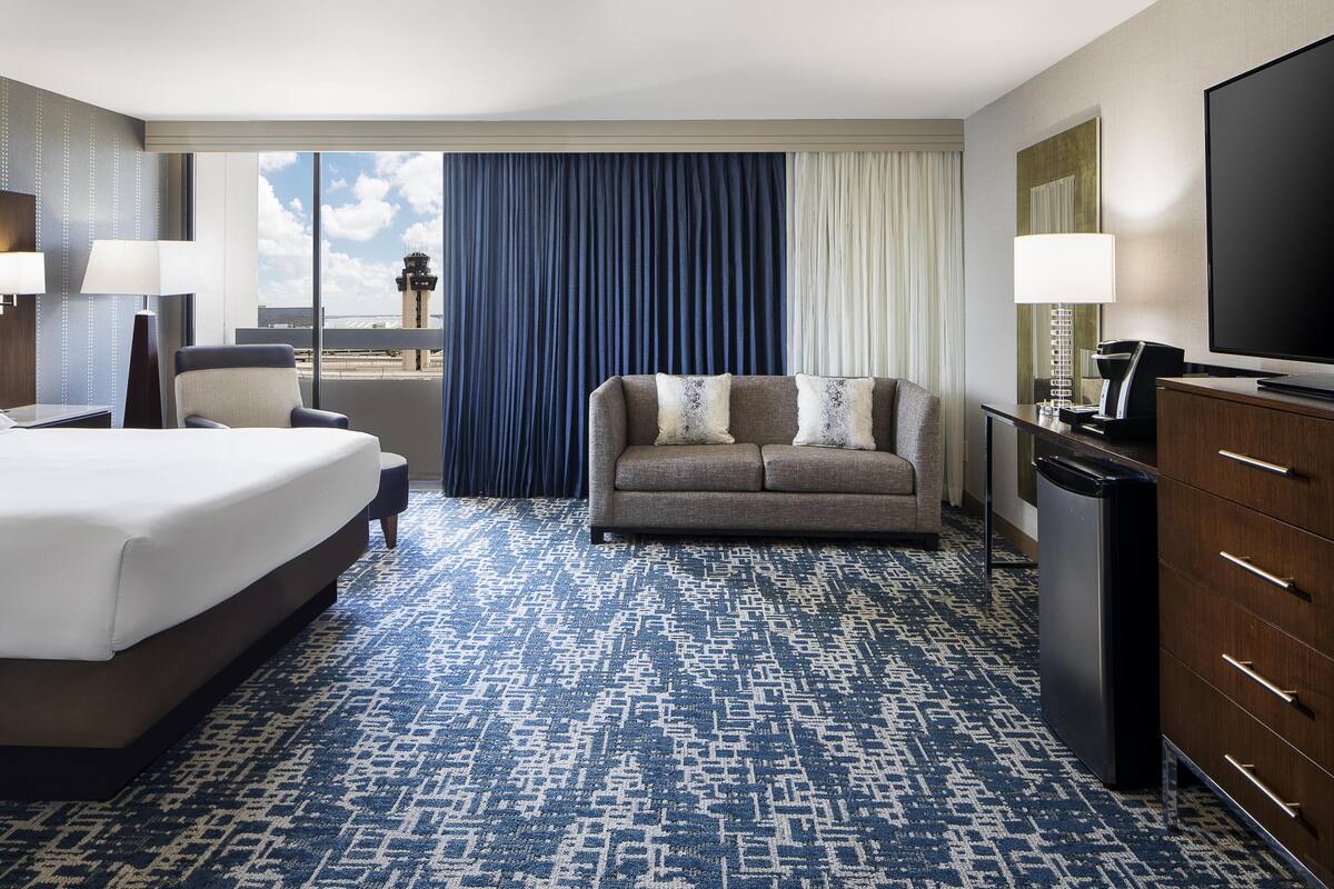 A hotel room with a bed, a sofa, a television, a desk with a lamp, and a view of the airport control tower.