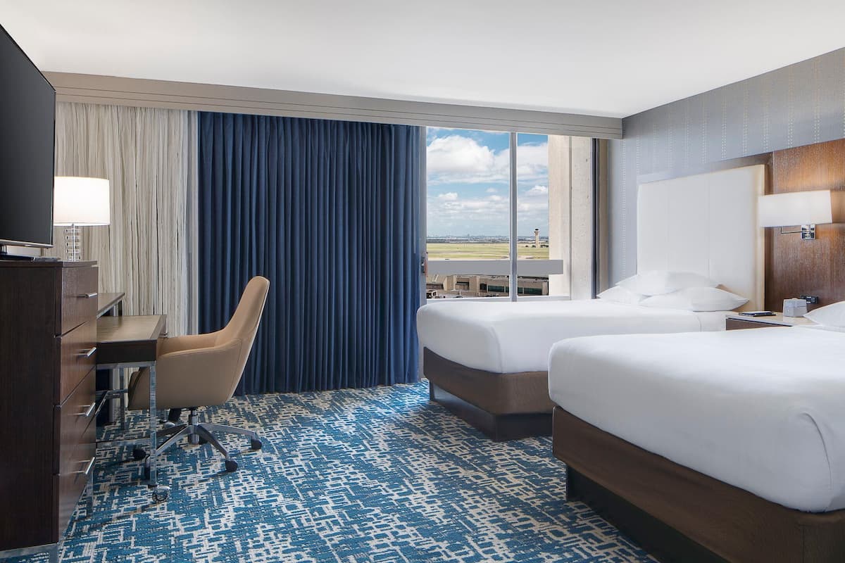 A hotel room with two beds, a desk, a chair, and a large window offering a view of the airport.