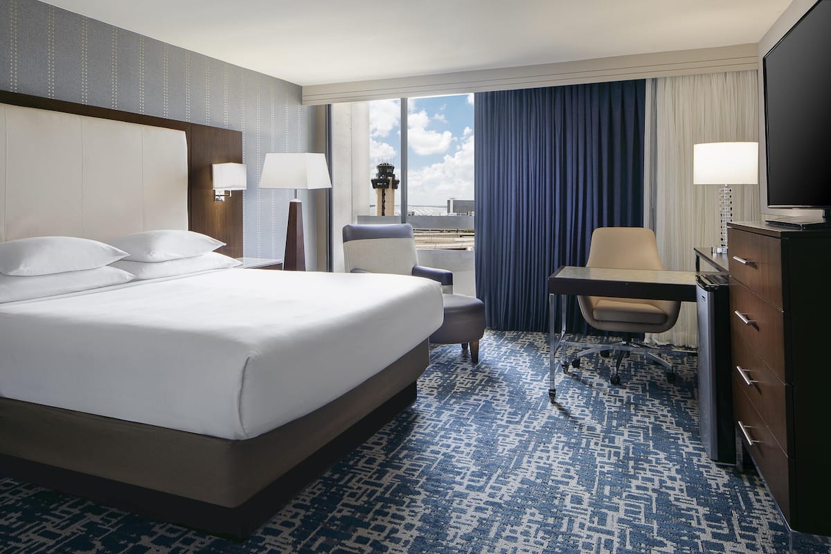 A hotel room with a large bed, a desk with a chair, a TV, and a view of the sky.