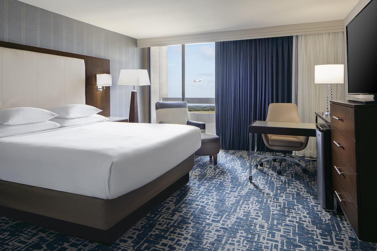 A hotel room with a large bed, a desk, a chair, a TV, and a view of an airplane outside the window.