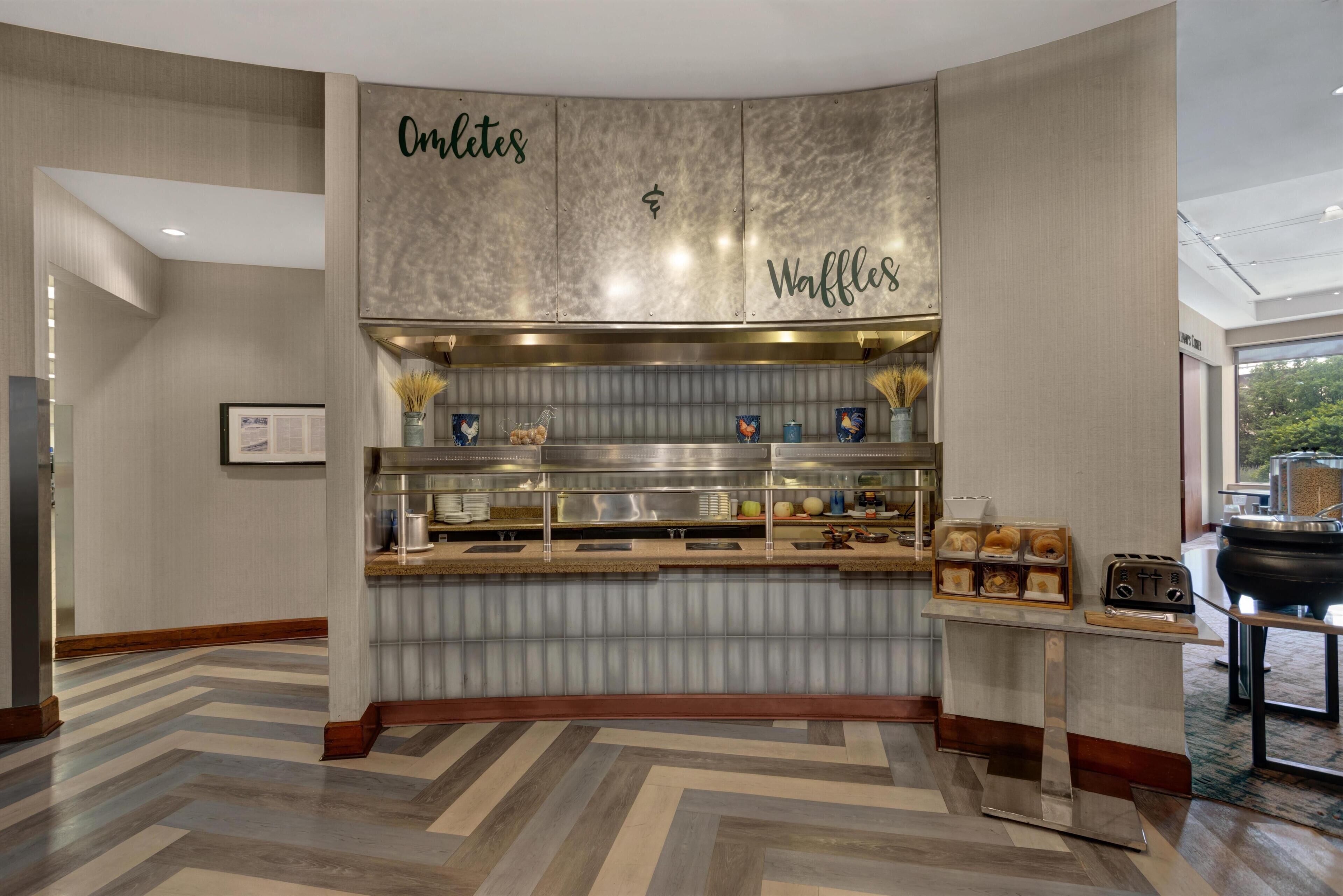 A breakfast bar with 'Omelettes' and 'Waffles' signage, a stainless steel refrigerator, and a buffet setup.
