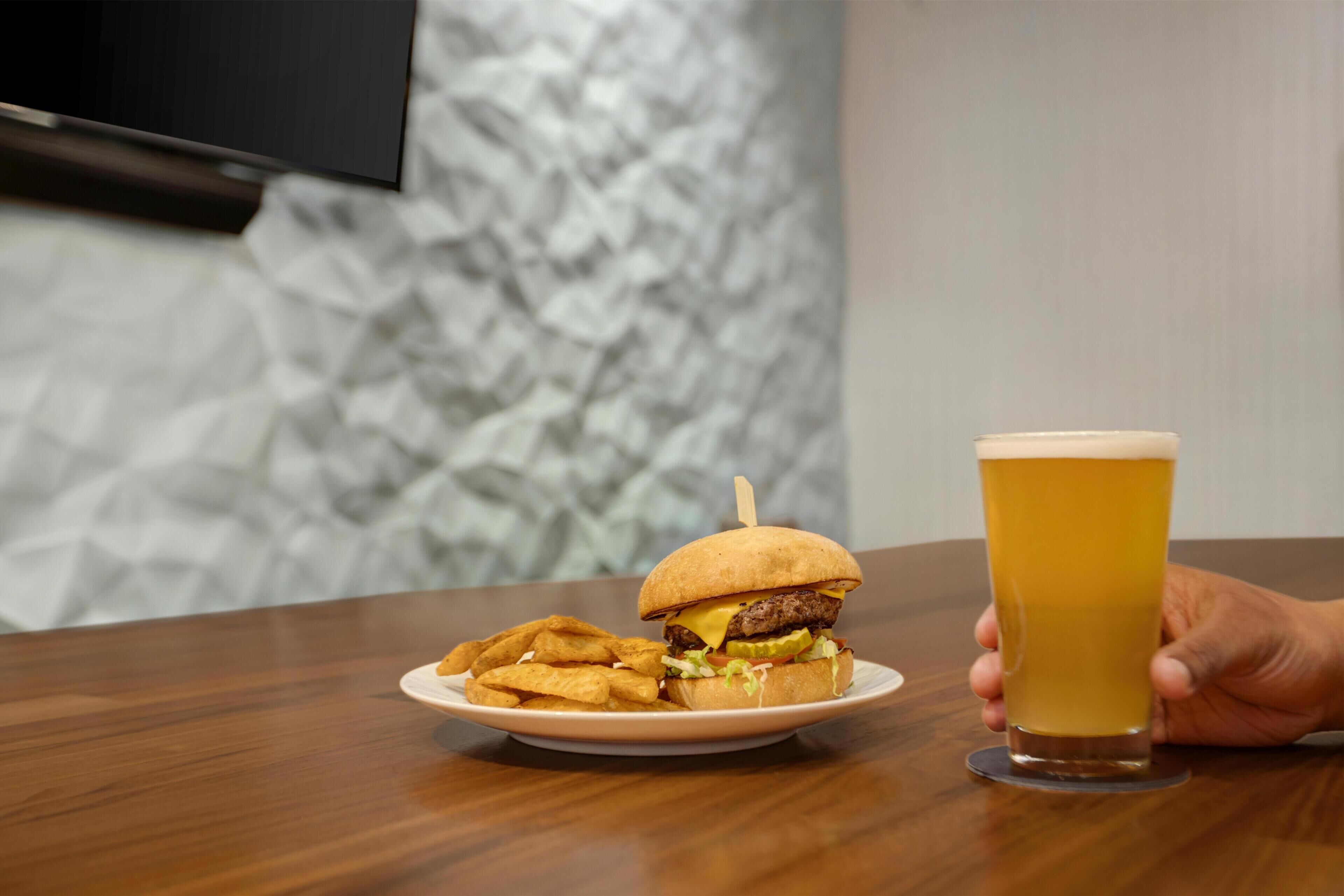 Un plato con una hamburguesa y papas fritas, una jarra de cerveza, y una mano sosteniendo la jarra.