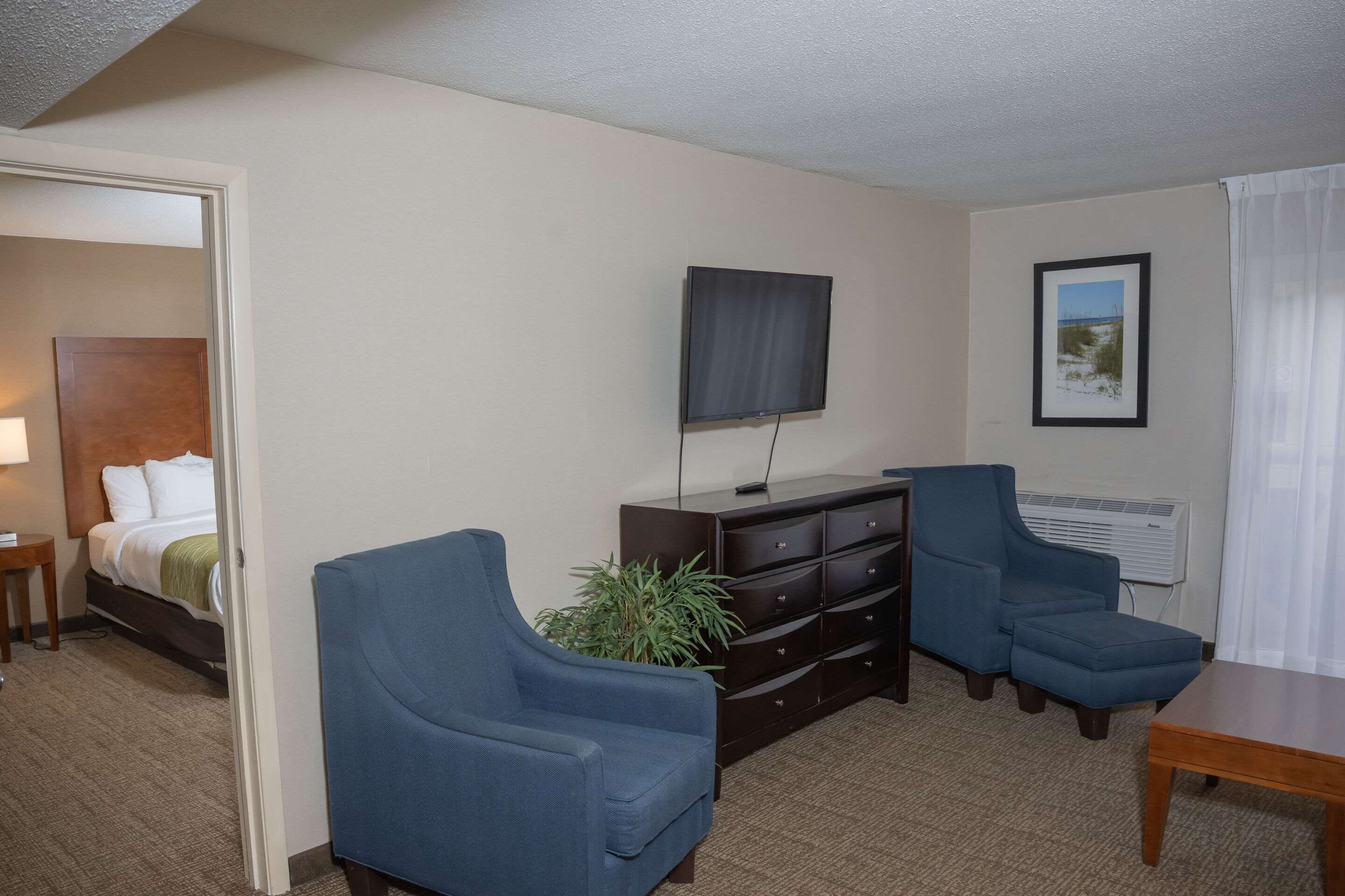 Una camera d'albergo con un letto, una scrivania, due poltrone blu, una TV, un comò e un quadro appeso al muro.