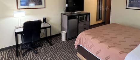 Habitación de hotel con cama, escritorio, silla, televisión, microondas y cubo de basura.