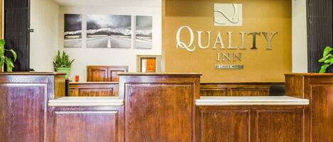 A reception area with wooden reception desks, a wall-mounted sign reading "Quality Inn by Choice Hotels," and three framed pictures on the wall.