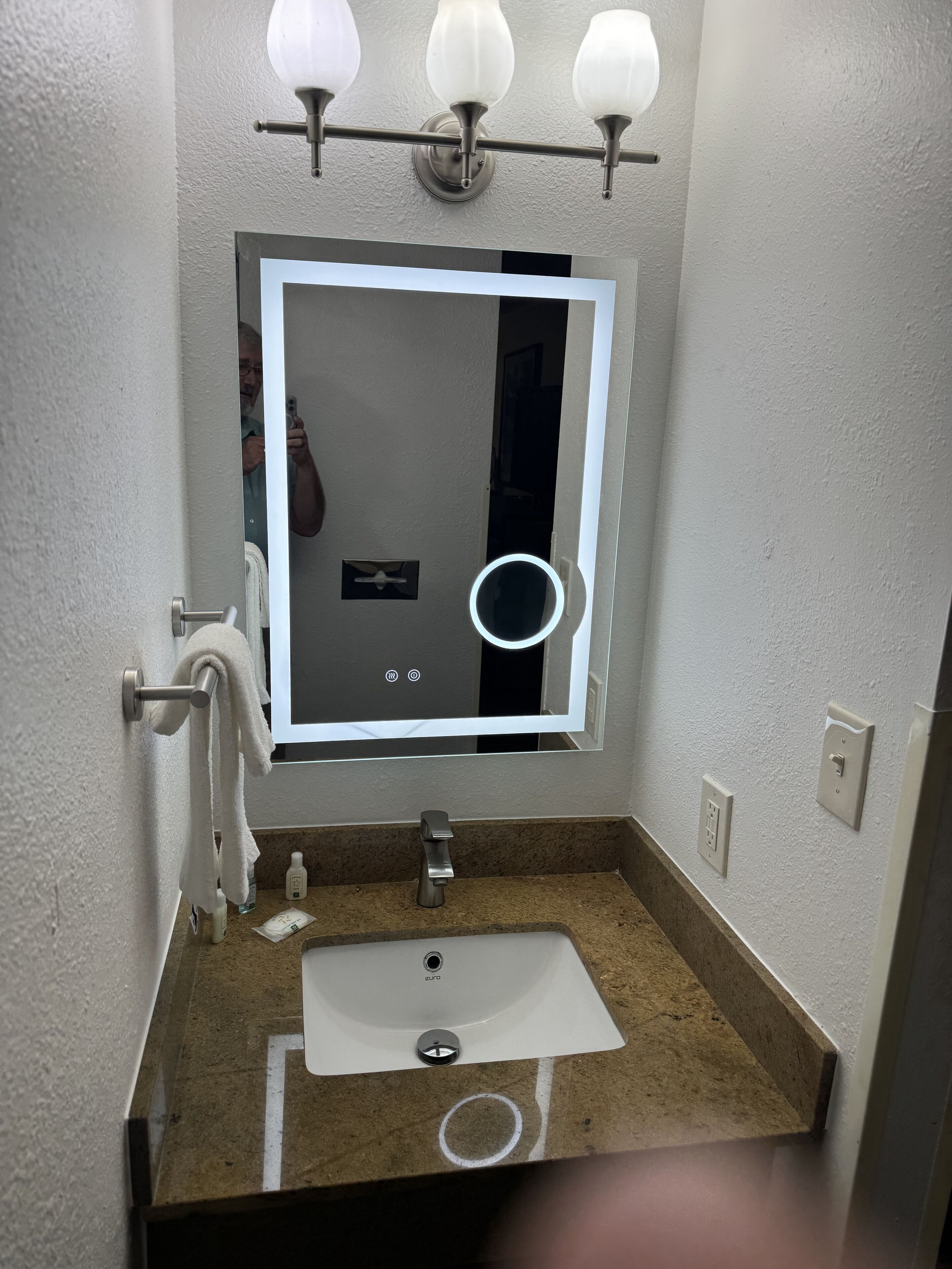 Une salle de bain avec un lavabo blanc, un miroir avec éclairage intégré, un porte-serviettes et un miroir de coiffeuse.