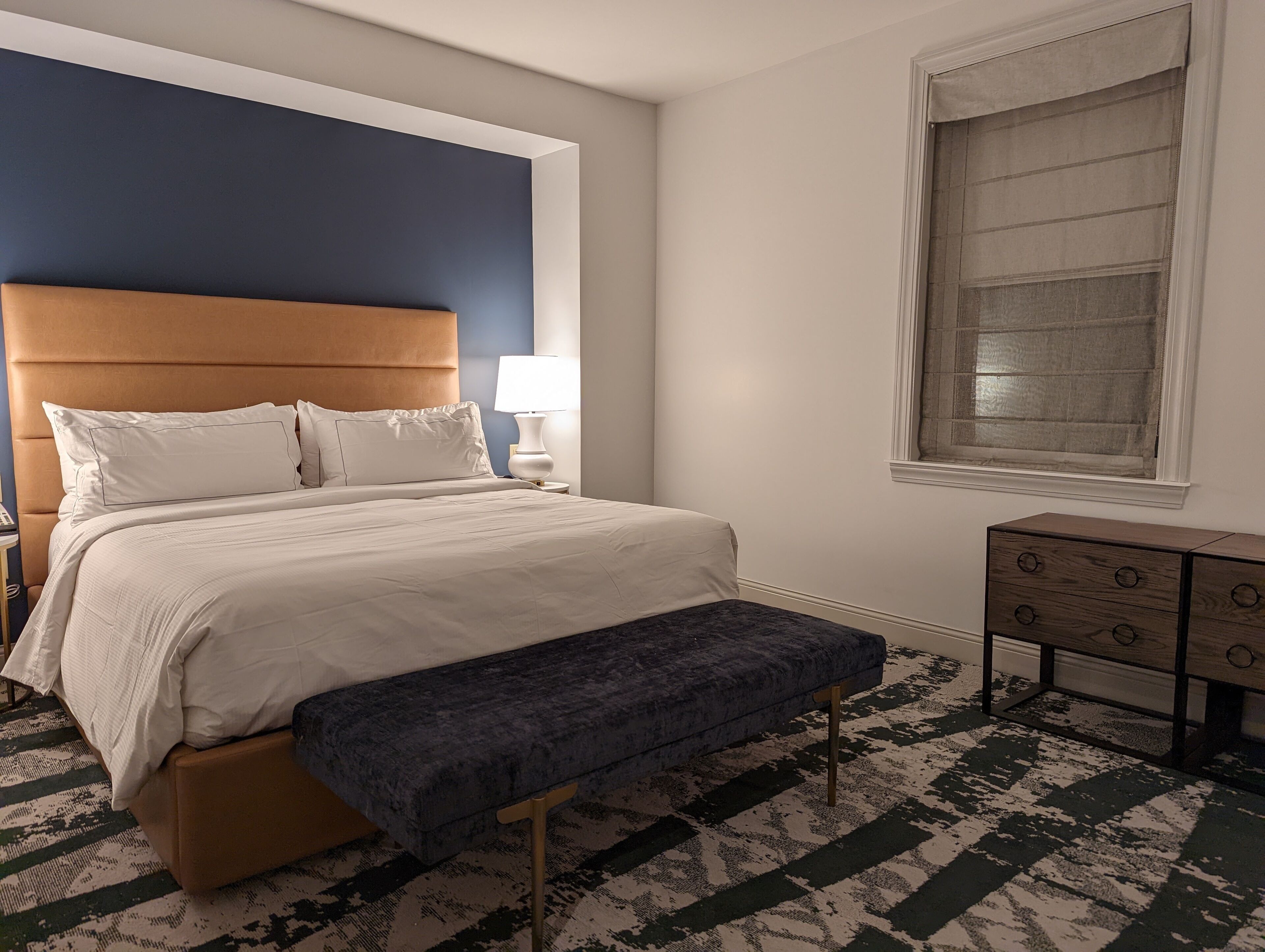 Un dormitorio ordenado con cama, banco, mesita de noche, lámpara y una ventana con cortinas.