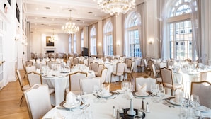 Un amplio salón de banquetes con varias mesas redondas, elegantes candelabros y grandes ventanales.