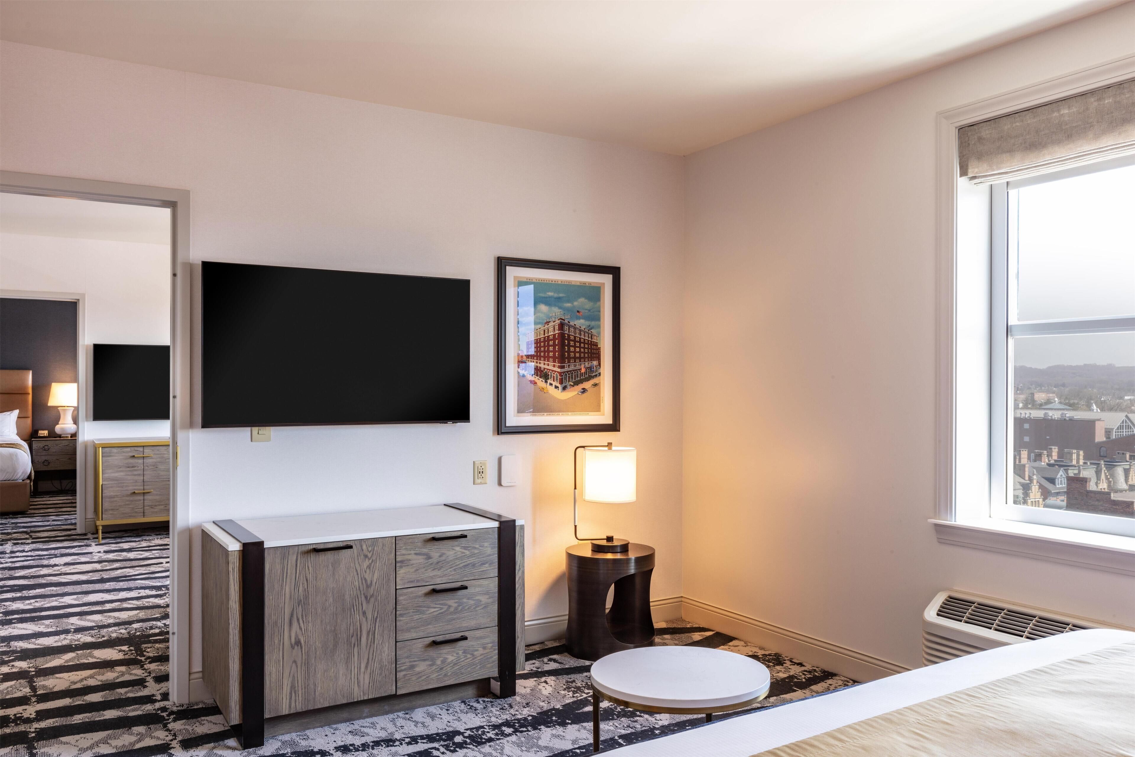 Habitación de hotel con televisión de pantalla plana, un cuadro enmarcado, lámpara de noche, una mesa redonda y vista a la ciudad desde la ventana.