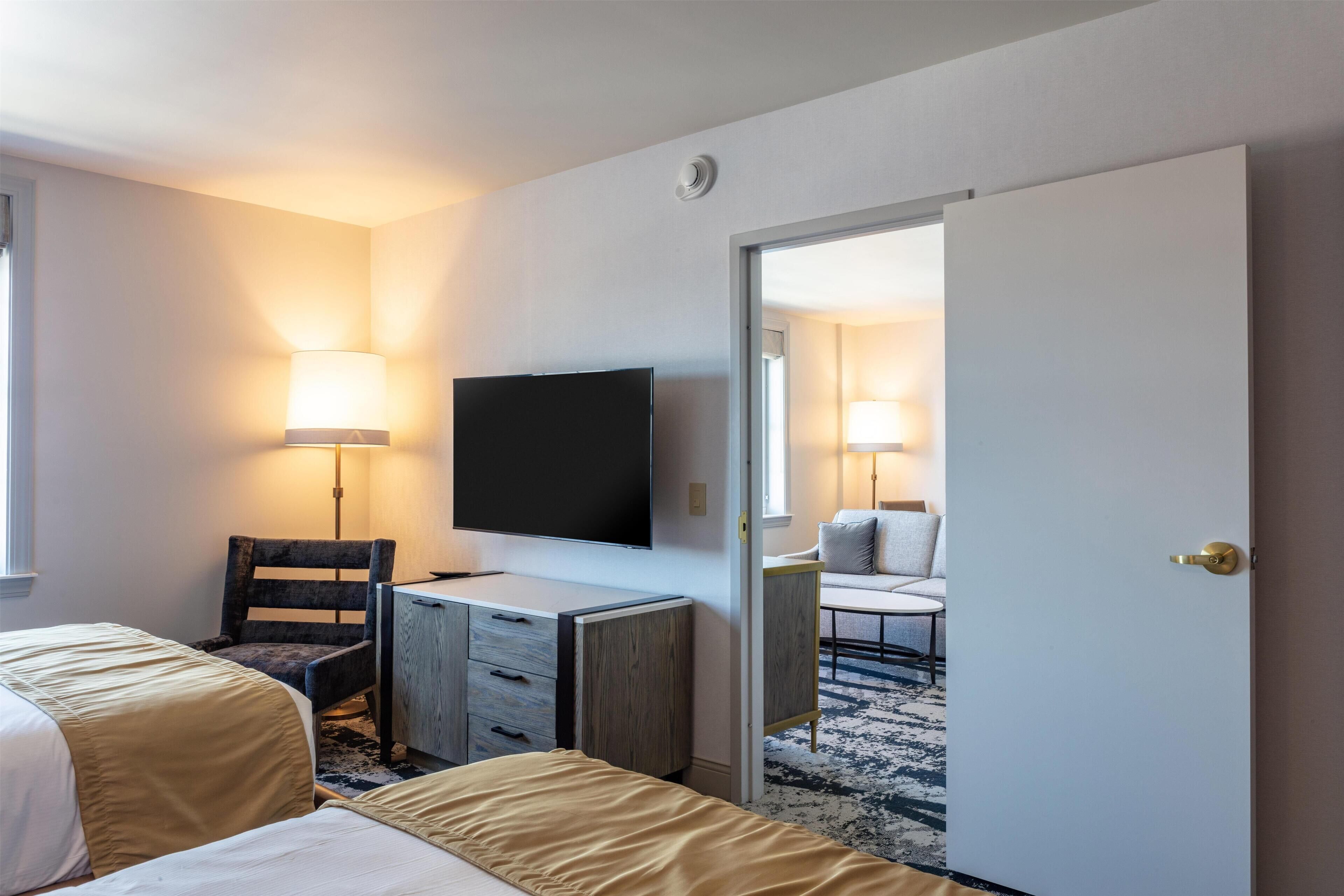 A hotel room with two beds, a desk, a chair, a TV, and a door leading to another room.