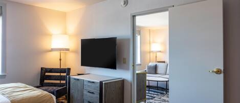Habitación de hotel con dos camas, un escritorio, una silla, un televisor y una puerta que da a otra habitación.