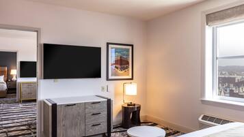 A hotel room with a flat-screen TV, a framed picture, a bedside lamp, a round table, and a window with a city view.
