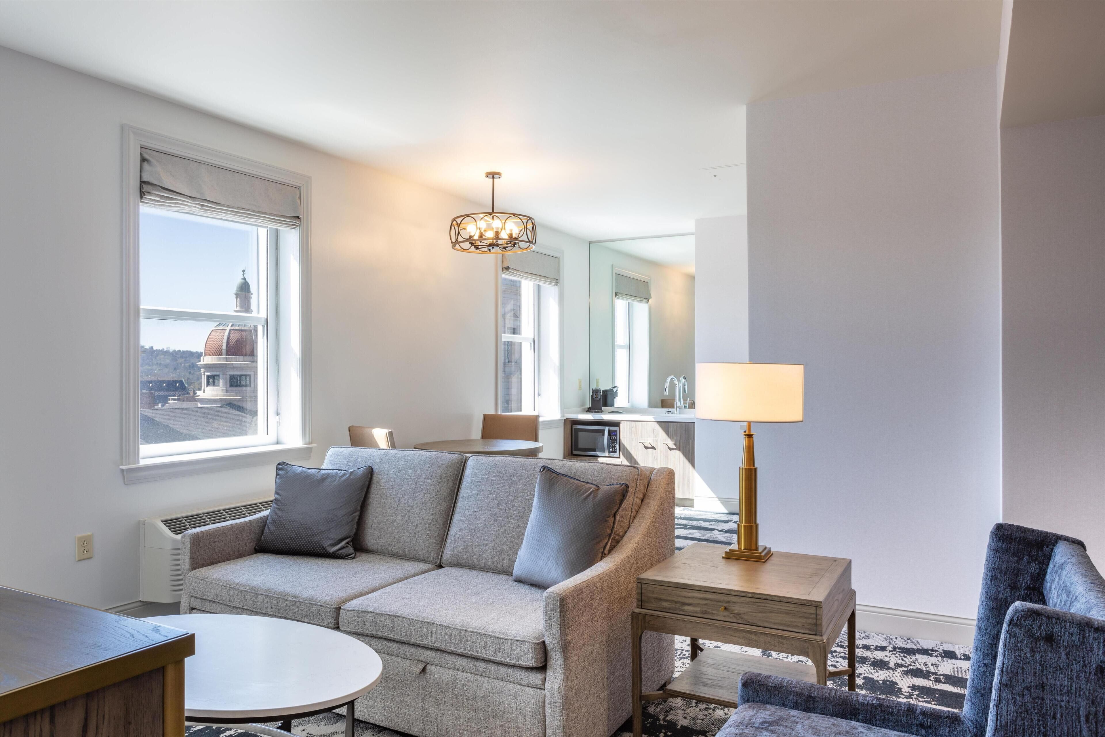 A modern hotel room with a sofa, a round table, a lamp, and a view of a building through the window.