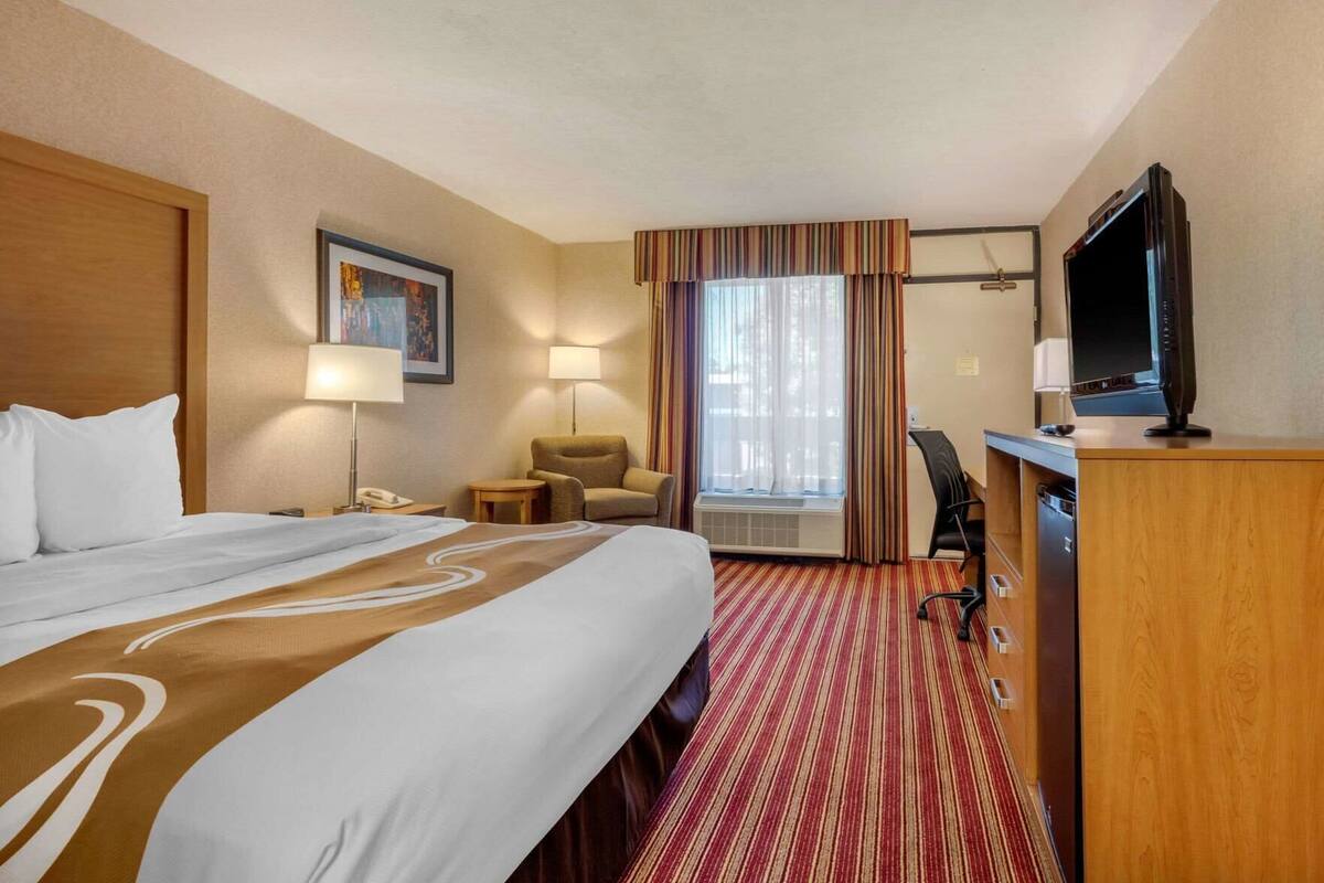 A hotel room with a large bed, a desk with a television, a chair, a lamp, and a window with curtains.