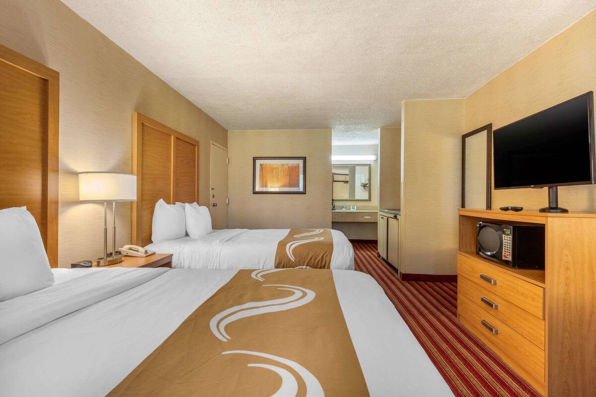 A hotel room with two beds, a TV, a microwave, a desk with a lamp, and a bathroom visible in the background.