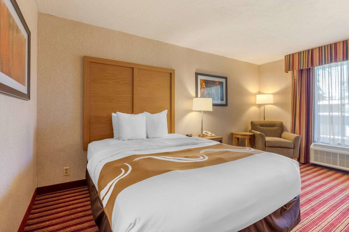 A hotel room with a large bed, a wooden headboard, a desk with a lamp, a chair, and a large window with curtains.