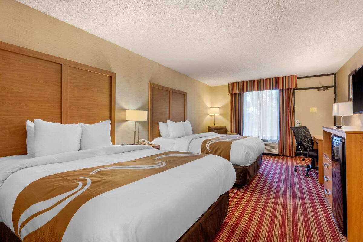 A hotel room with two beds, a desk, a chair, a TV, and a window with curtains.