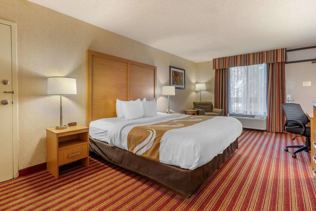 A hotel room with a large bed, a desk with a computer, a chair, a lamp, and a window with curtains.