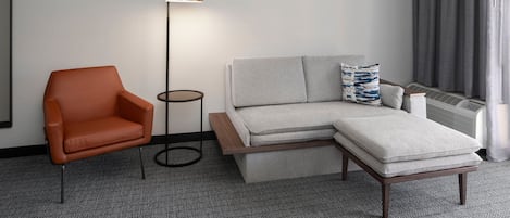 Una habitación de hotel moderna con un sofá gris, un sillón naranja, una mesita auxiliar y una lámpara de pie.