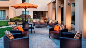 An outdoor patio with wicker furniture and an umbrella, surrounded by a well-maintained garden and a building facade. - Courtyard by Marriott Little Rock West (Little Rock)