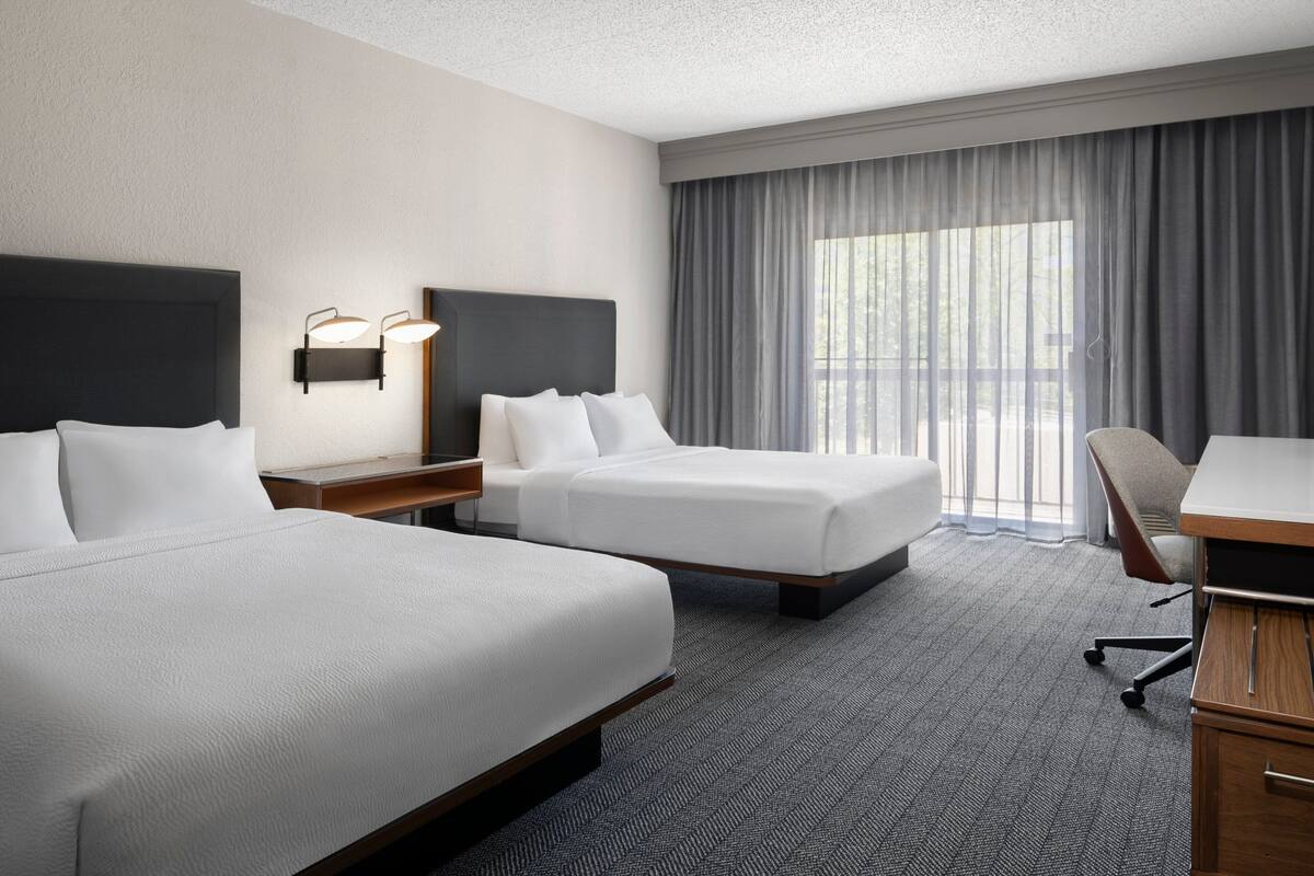 Een hotelkamer met twee bedden, een bureau, een stoel en een groot raam met gordijnen.